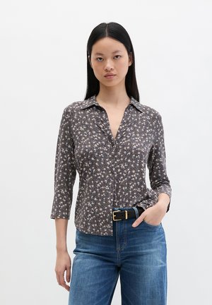 Woman with straight black hair wears a brown floral button-up shirt, blue jeans, and a black belt with a gold buckle, hand in pocket.