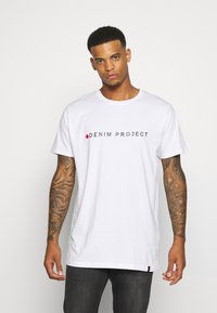 Balta medvilninė marškinėliai su juodu ir raudonu spaudu "DENIM PROJECT" logotipu per krūtinę. Trumpi rankovės, laisvas siluetas, minimalus dekoravimas.