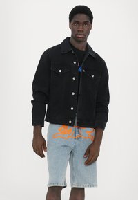 Veste en denim noir avec fermeture à boutons, deux poches poitrine et une coupe ajustée, associée à un short bleu clair avec un graphique orange.