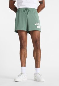 Mesh idrettsshorts i lys grønn med elastisk midjebånd, front snøring og hvit trykt logo. Kombinert med hvite joggesko og sokker.