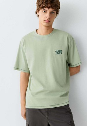 Jonge man met krullend haar, die een lichtgroen T-shirt draagt met de tekst "JAPAN" op de borst en een donkere broek, staand tegen een effen witte muur.
