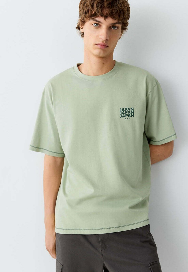 Jeune homme aux cheveux bouclés portant un t-shirt vert clair avec le texte « JAPAN » sur la poitrine et un pantalon sombre, debout contre un mur blanc uni.