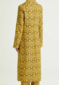 Lungo cappotto giallo con un motivo geometrico di ovali e cerchi intrecciati, realizzato in tessuto liscio con una silhouette strutturata.
