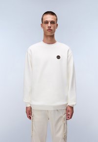White sweatshirt with a round neckline, ribbed cuffs, and a small black and orange patch on the chest, paired with beige cargo pants.