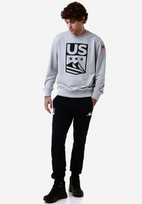 Felpa grigia con logo americano nero e grafica di montagne, abbinata a pantaloni joggers neri con dettagli bianchi e stivali neri.