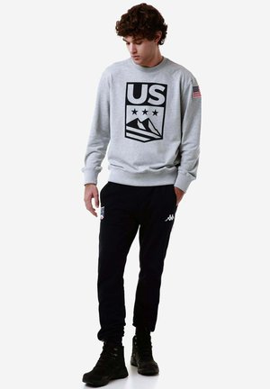 Felpa grigia con logo americano nero e grafica di montagne, abbinata a pantaloni joggers neri con dettagli bianchi e stivali neri.