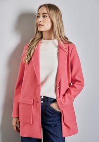 Cappotto in misto lana rosa con revers e tasche frontali, abbinato a una camicia bianca e jeans scuri. Texture liscia, design strutturato.