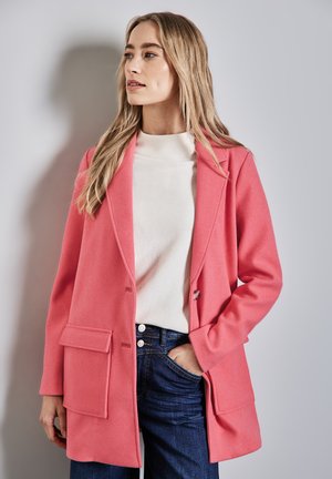 Abrigo de mezcla de lana rosa con solapas y bolsillos frontales, combinado con una camisa blanca y jeans oscuros. Textura suave, diseño estructurado.