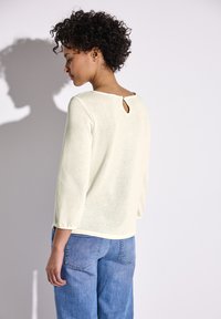 Maglione giallo chiaro, con texture e maniche a tre quarti, con un piccolo dettaglio a chiave sulla schiena, abbinato a jeans in denim blu.