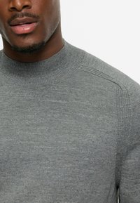 Grauer gestrickter Pullover mit hohem Halsausschnitt, der eine glatte Textur und subtile Variationen in der Garnfarbe aufweist, entworfen für eine figurnahe Passform.