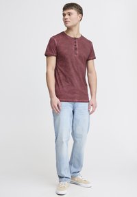 Burgunderfarbenes Kurzarm-Henley-T-Shirt mit drei Knöpfen, kombiniert mit hellblauen Jeans und beigen Sneakers. Der Hintergrund ist weiß.