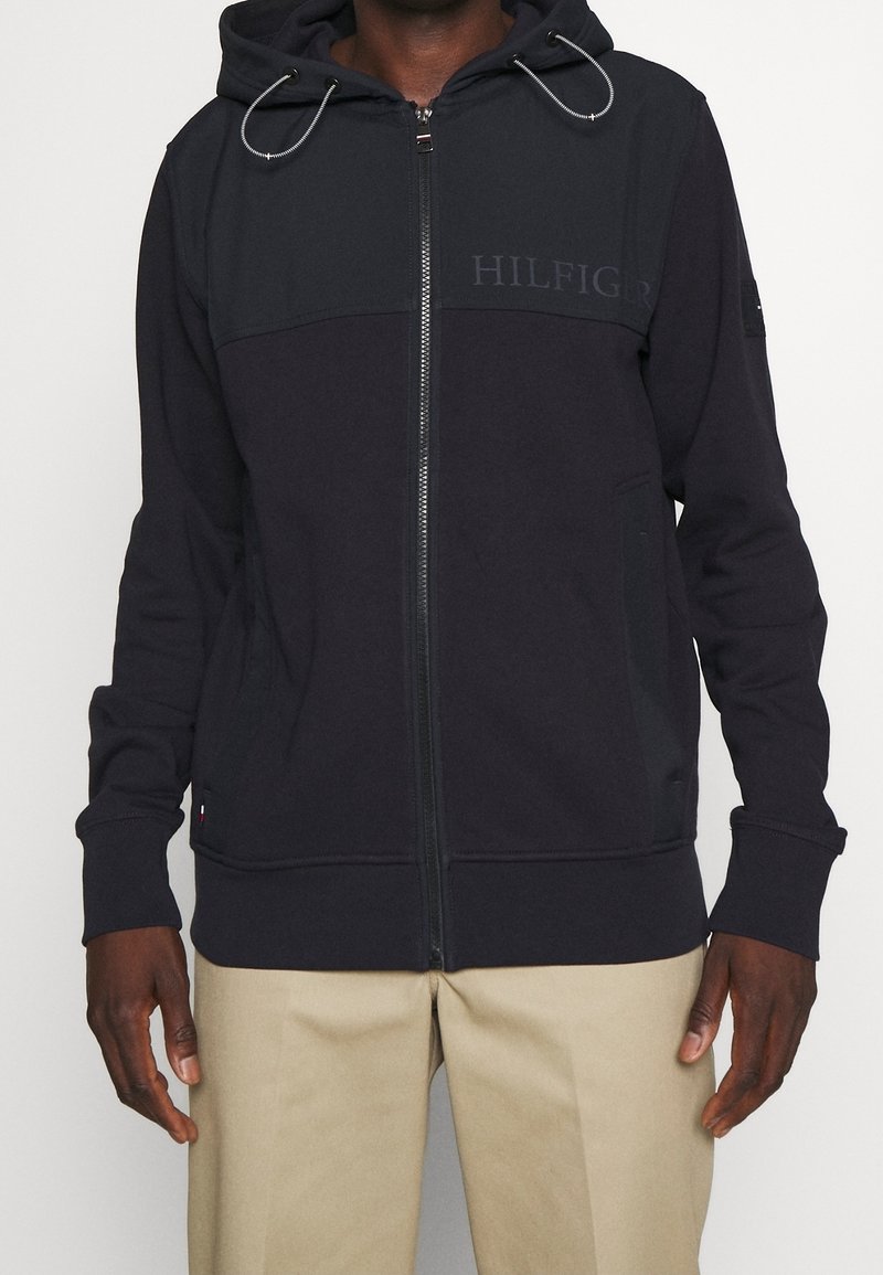 Sudadera con capucha y cremallera en azul marino oscuro, presenta cierre de cremallera, cordón ajustable en la capucha y un logo en el pecho. Tejido texturizado.