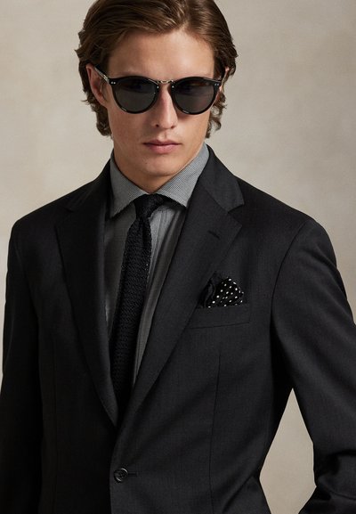 Junger Mann trägt schwarzen Anzug, gemustertes Hemd, gestrickte Krawatte, schwarze Sonnenbrille und gepunktetes Einstecktuch vor neutralem Hintergrund.