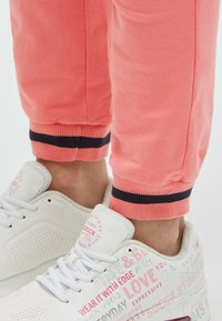 Korallfarbene Jogginghosen mit schwarzen gestreiften Bündchen, kombiniert mit weißen Turnschuhen, die Text- und Grafikakzente in Pink und Grau zeigen. Weiche Stofftextur.
