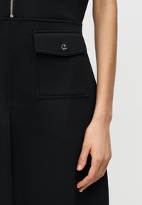 Close-up of a black garment with a large flap pocket featuring a round metallic button, alongside a relaxed right hand.