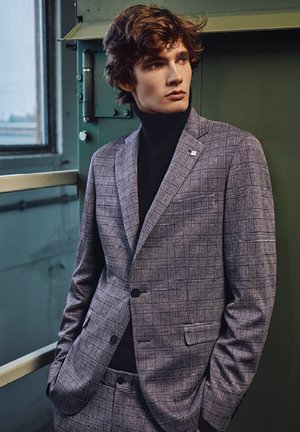 Gray plaid blazer with a notch lapel, single button, and inset pocket. Worn over a black turtleneck. Smooth, tailored fit.