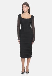 Femme portant une robe midi noire ajustée avec des détails froncés, des manches longues transparentes, un décolleté carré et des escarpins noirs à talons aiguilles.