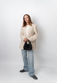 Femme debout sur fond blanc, portant un blazer beige, un haut blanc, un jean bleu clair, des chaussures noires, tenant un petit sac à main noir par les poignées.