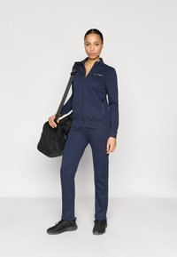 Αθλητική φόρμα ναυτικού μπλε από μαλακό ύφασμα, με ζακέτα φερμουάρ και τσέπες, και αντίστοιχο παντελόνι, συνοδευόμενα από μια μαύρη τσάντα duffel.