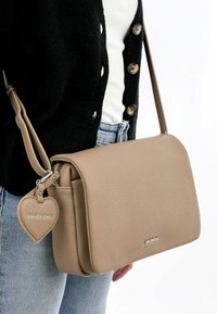 Sac bandoulière en simili cuir beige avec un rabat, surface texturée et une étiquette en forme de cœur. Le sac se ferme avec une fermeture éclair et dispose d'une sangle réglable.