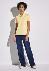 Gul polo skjorta med klassisk krage, korta ärmar, parat med mörkblå utsvängda jeans och vita sneakers, framför en neutral bakgrund.