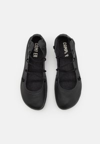 Paire de ballerines lacées noires Camper avec bouts arrondis et semelles en tissu visibles de dessus sur fond blanc.