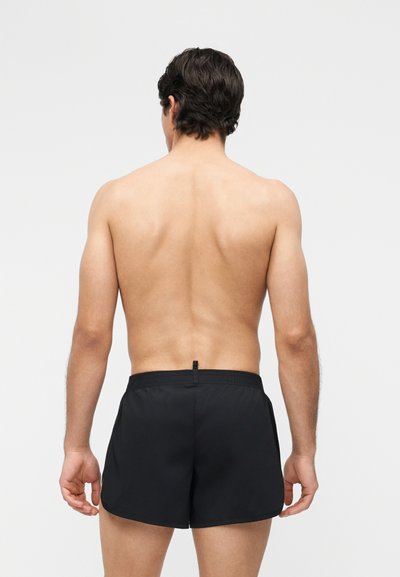 Άνδρας με σκούρα μαλλιά στέκεται γυμνός από τη μέση και πάνω, γυρισμένος πλάτη, φορώντας μαύρο μαγιό μπροστά σε απλό λευκό φόντο.