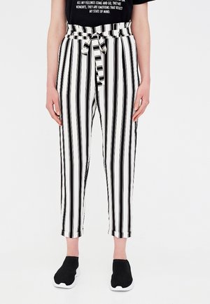 Persona que lleva pantalones de talle alto con rayas verticales en blanco y negro, cinturón, camiseta negra y zapatos negros sin cordones.