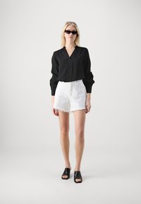Schwarze Bluse mit langen Ärmeln und V-Ausschnitt, kombiniert mit weißen Shorts mit fransigem Saum und schwarzen offenen Mules. Einfaches, modernes Design.