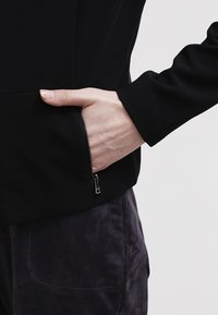 Main partiellement dans la poche zippée d'une veste noire portée sur un pantalon sombre et texturé.