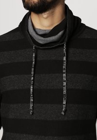 Czarna i szara bluza z kapturem w paski, z golfem, teksturowaną tkaniną i kontrastową warstwą wewnętrzną. Wiązania przy szyi dodają szczegółów.