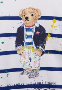 Camiseta con una ilustración de un oso que lleva un blazer azul marino, una camisa a rayas y unos pantalones vaqueros pintados. Salpicaduras brillantes sobre una tela blanca, con rayas azul marino en la parte inferior.