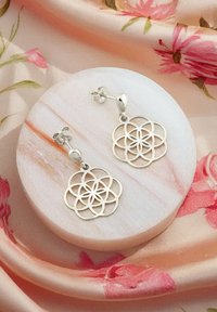 Orecchini d'argento con un design floreale e geometrico ritagliato. Presentano una chiusura a perno e sono esposti su un supporto rotondo e marmorizzato.