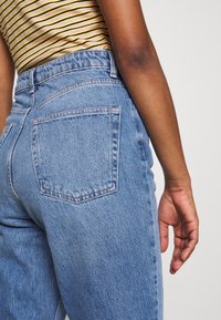 Persona con jeans de mezclilla azul de talle alto y una camisa amarilla a rayas, con el brazo derecho colgando relajado a su lado.