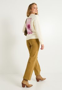 Un pull crème avec un design graphique rose dans le dos, associé à un pantalon large moutarde et des bottines à talons beiges.