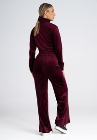 Burgundowy dres z weluru z wysokim kołnierzem, dopasowanymi długimi rękawami oraz spodniami o szerokich nogawkach z elastycznym pasem. Białe sneakersy z falistą krawędzią.