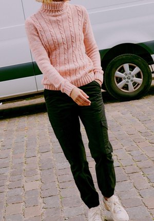 Person wearing a pink cable-knit sweater, black cargo pants, and white sneakers standing on cobblestone near a white vehicle.