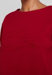 Red cotton sweatshirt featuring an embossed crocodile logo, round neckline, and smooth texture, emphasizing a minimalist design.