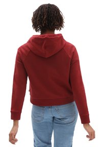 Felpa rossa con cappuccio, tasca frontale, polsini a costine e cordoncino. Indossata con jeans di denim blu chiaro. Tessuto morbido e texture.