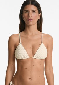 Bikini top a triangolo di colore crema con tessuto testurizzato, spalline sottili e un dettaglio dorato al centro.