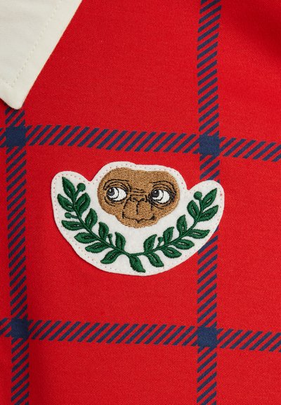 Κόκκινο ύφασμα με καρώ σχέδιο σε ναυτικό μπλε. Περιέχει μια κεντητή στάμπα με το πρόσωπο ενός χαρακτή�ρα περιτριγυρισμένο από πράσινα φύλλα σε λευκό φόντο.