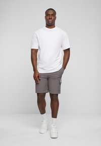 T-shirt blanc à manches courtes, short cargo gris avec poche latérale, baskets blanches et chaussettes blanches, se tenant sur un fond neutre.