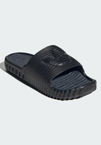 Svart slide-sandal med en texturerad ovansida som har horisontella ränder och en logotyp. Grå fotbädd med små, upphöjda prickar för grepp.