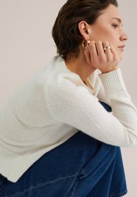 Vit stickad sweater med rundad krage och ribbade ärmar. Modellen har på sig mörkblå jeans, med händerna vilande på knäna, vilket visar upp minimalistisch guldsmide.