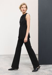 Blazer senza maniche nera con dettagli dei bottoni, abbinato a pantaloni larghi neri. La modella si trova su un pavimento testurizzato contro uno sfondo neutro.