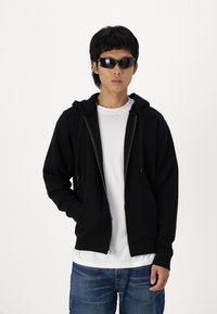 Zwarte rits hoodie met een voorzak, gecombineerd met een wit t-shirt en blauwe jeans. Model draagt zwarte zonnebril.