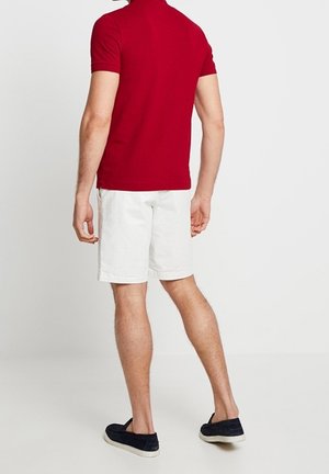 Polo rouge à manches courtes, porté avec un short blanc. La tenue est complétée par des chaussures sans lacets bleu foncé. Design propre et simple.