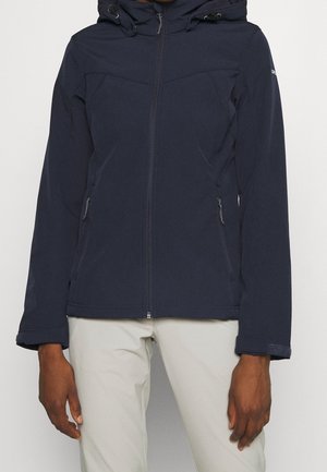 Personne portant une veste zippée bleu marine avec capuche et un pantalon beige clair, debout devant un fond clair uni.