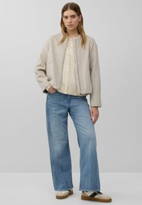 Lichtbeige bomberjack met een rits aan de voorkant, gecombineerd met een crèmekleurig overhemd met knopen en lichtblauwe brede jeans. Sneakers hebben beige en zwarte accenten.