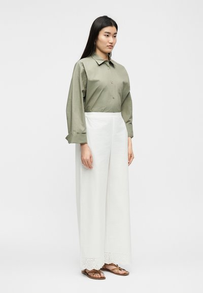 Γυναίκα όρθια με πουκάμισο σε ελιά πράσινο κουμπωτό, λευκό παντελόνι φαρδύ με κέντημα στο τελείωμα και καφέ σανδάλια μπροστά σε απλό φόντο.